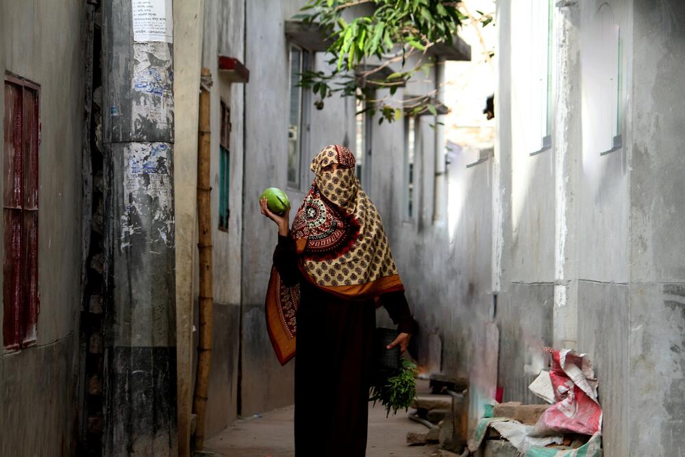 Photo of a lady wearing traditional clothes covering also her head and face, in an alley, holding a papaya in one hand