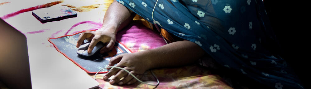 An adolescent using the computer on her bed with her phone next to it. The adolescent's face is not in the picture.
