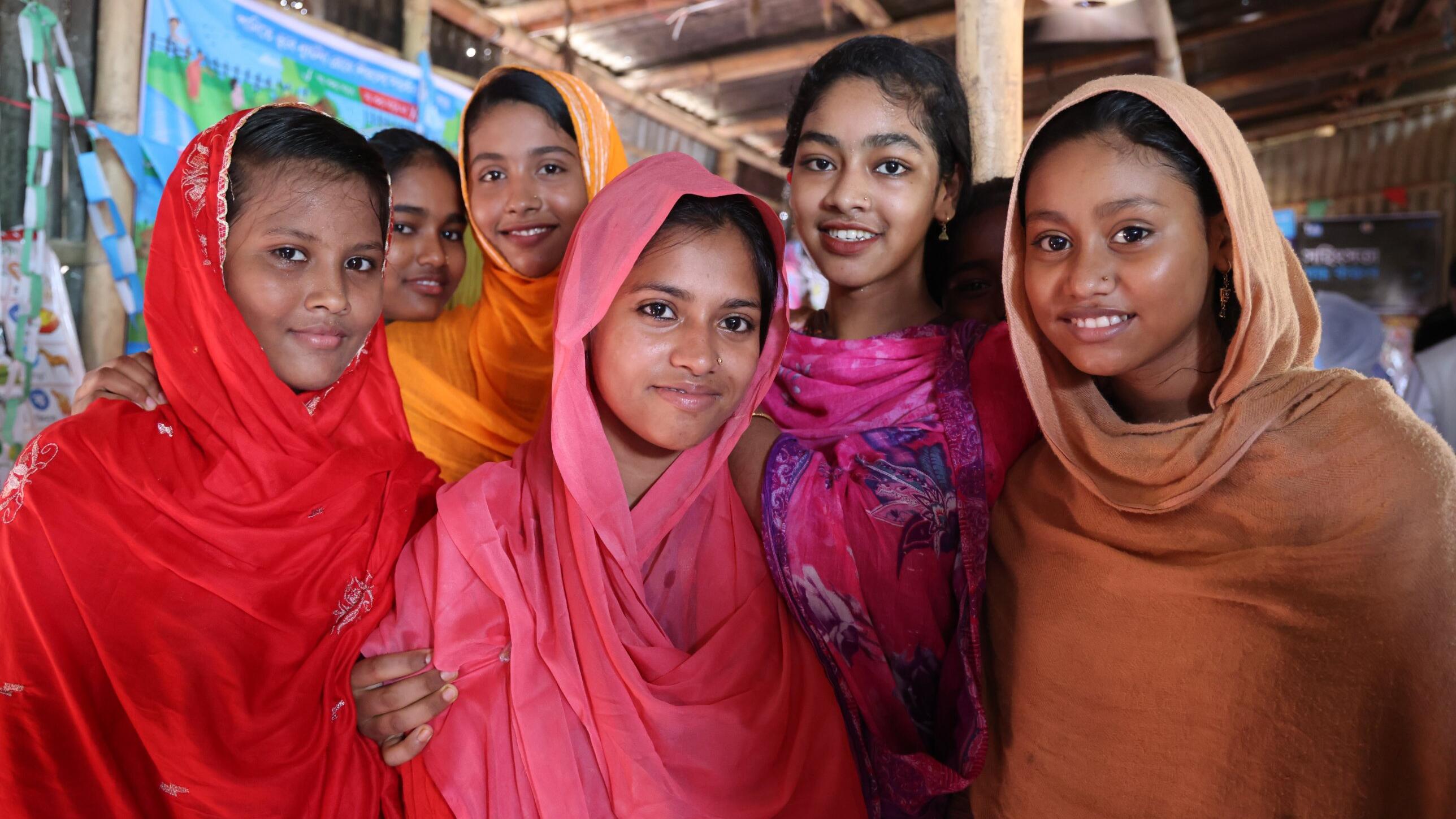 Five smiling adolescent girls in colourful headscarves of red, pink, orange and brown stand close together inside a bamboo-and-tin learning space, with educational posters on the wall behind them.
