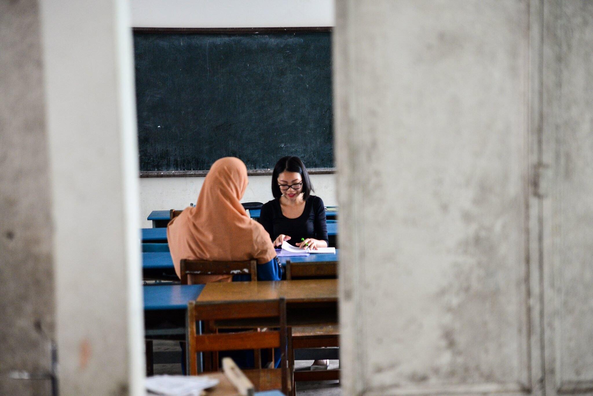 Two young women sitting in a classroom, one facing back wearing a scarf over her head and the other wears glasses