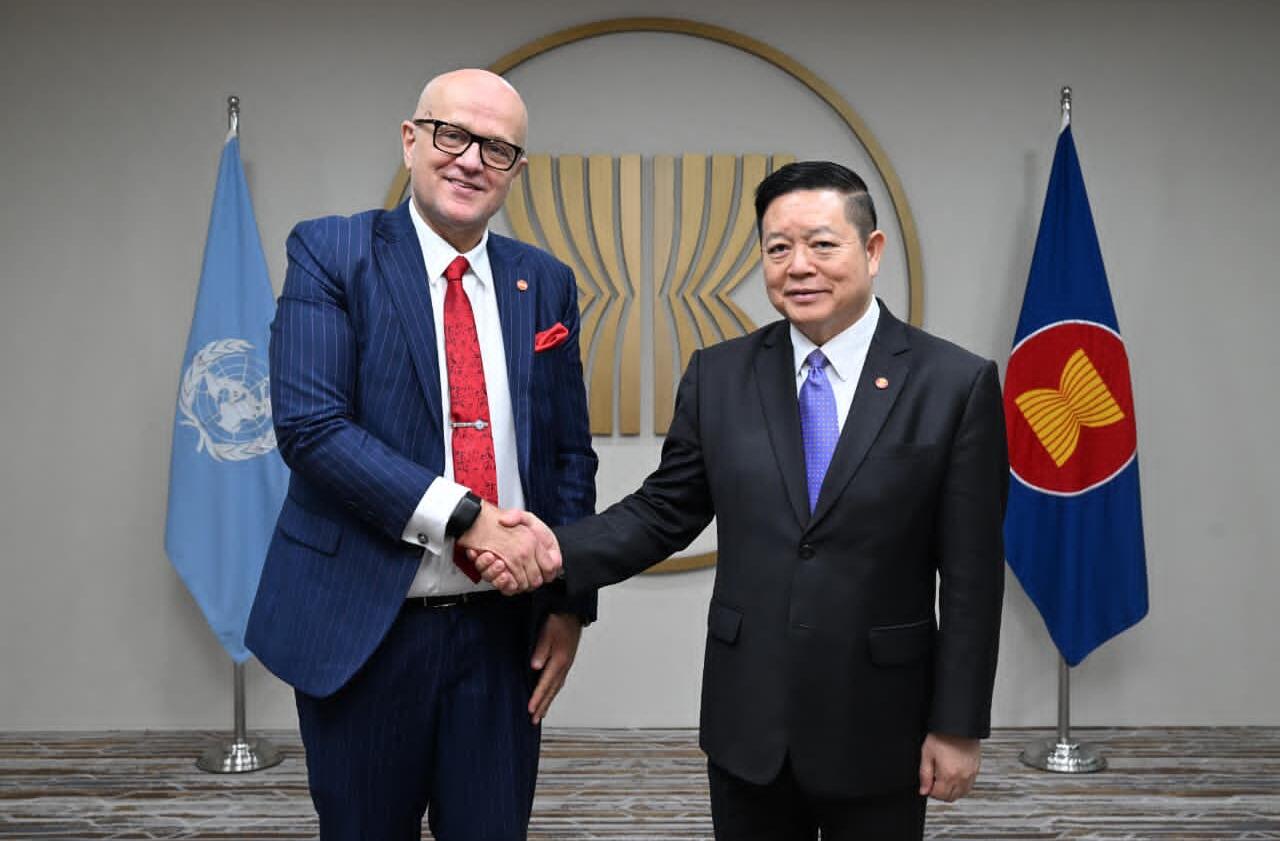 Two men wearing suits and shaking hands, with the UN and ASEAN flags in the background