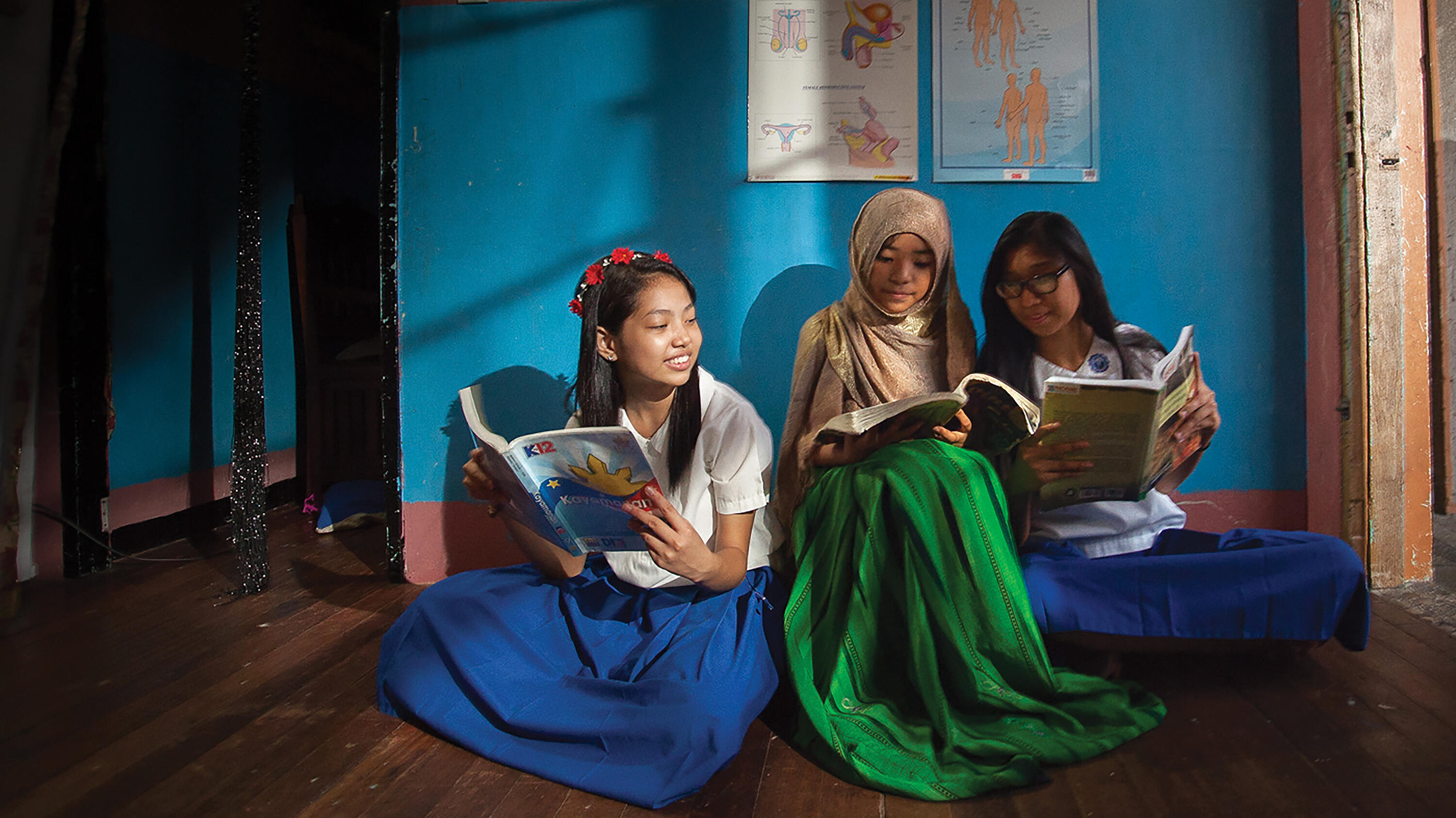 Three girls wearing school uniform, sitting on a wooden floor and reading school books